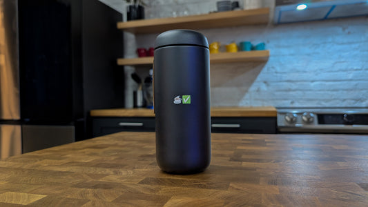 A black travel mug with a green coffee check logo, placed on a wooden surface, with kitchen items blurred in the background.